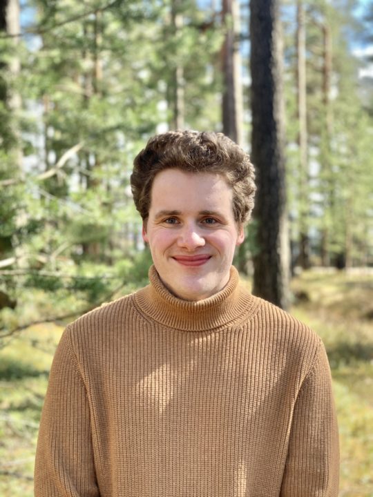 Fotografi. Portrettbilde av Henrik Aasgaard Sortland ikledd brun genser stående i skogen.