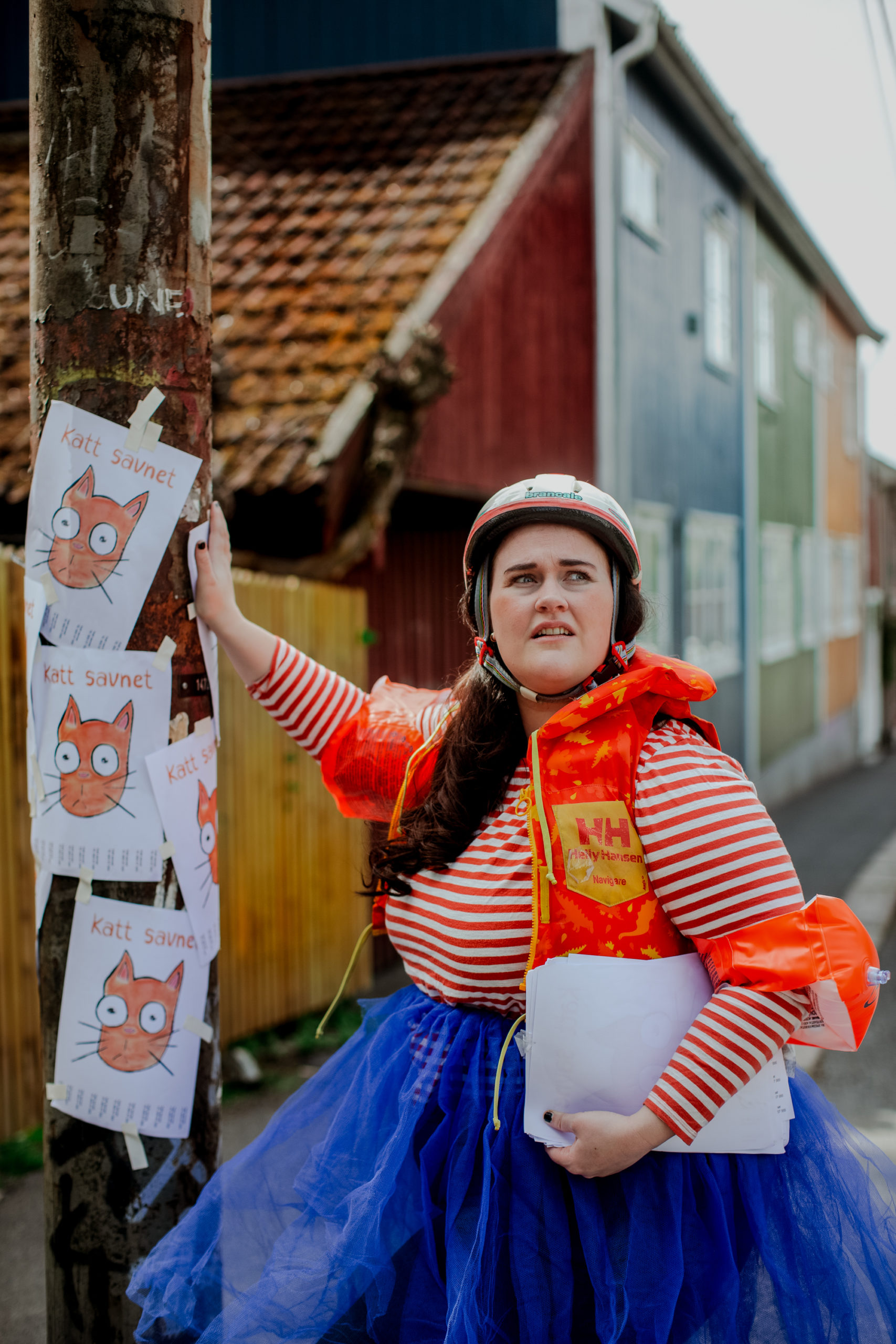 Fotografi: Bildet viser en ung, kvinnelig skuespiller som henger opp plakater av en savnet katt. Kvinnen har på seg oransje redningsvest, sykkelhjelm og oppblåsbare armringer.