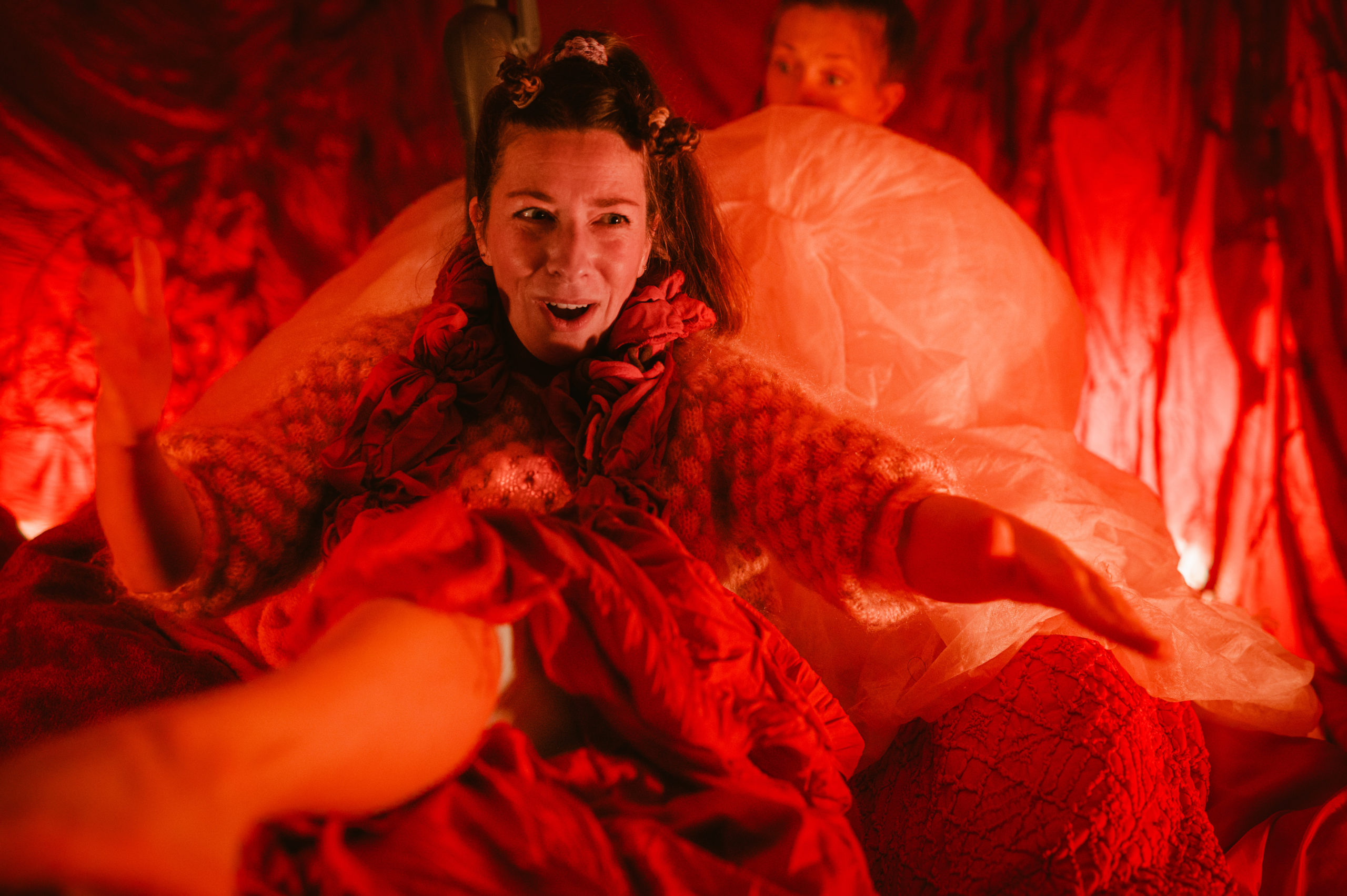 Fotografi: Bildet viser en kvinne med musefletter som har på seg røde klær. Hele rommet hun sitter er belyst i rødt og i bakgrunnen skimtes en annen kvinne.