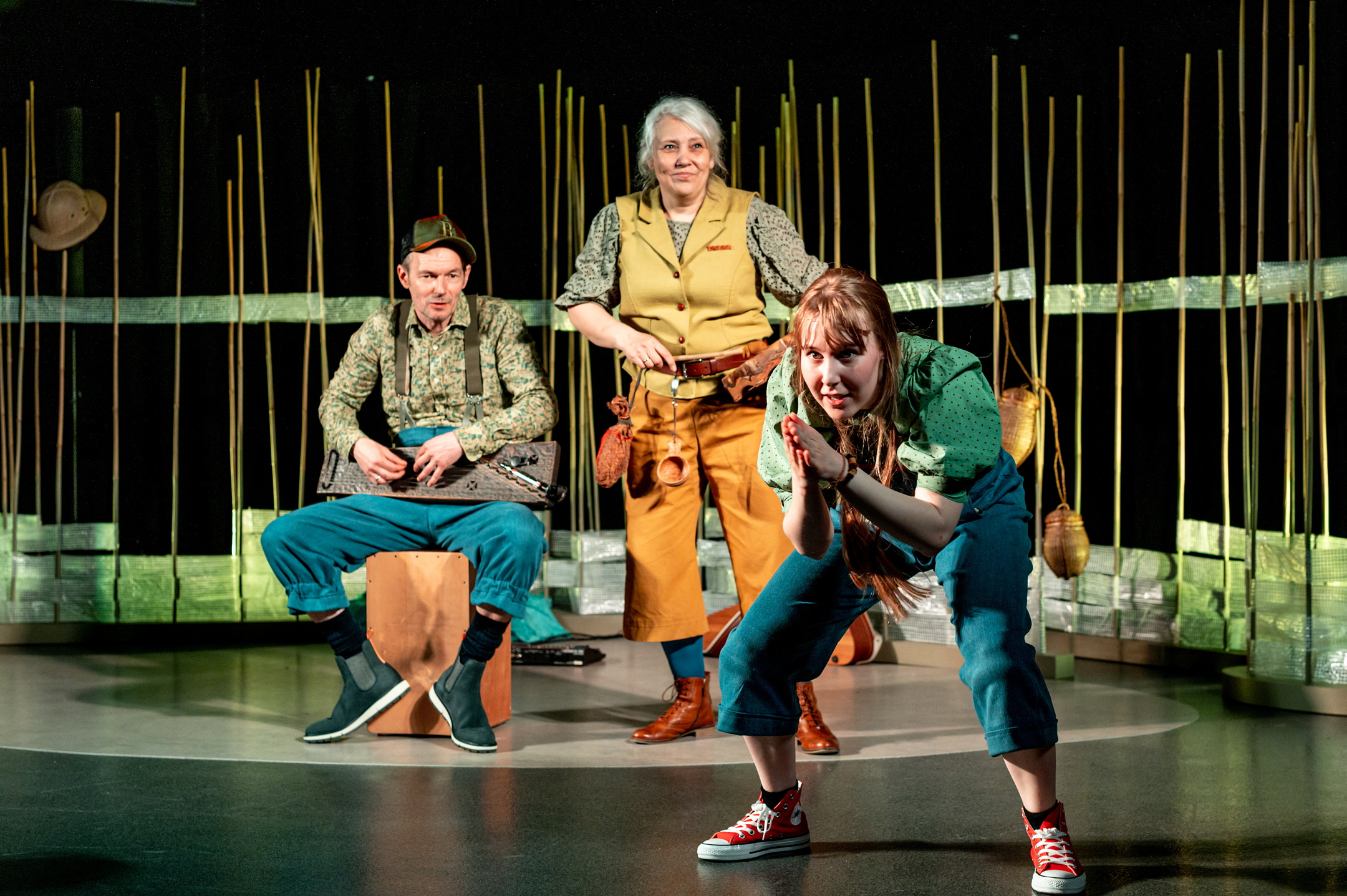 Fotografi: Bildet viser tre skuespillere på en scene, to kvinner og en mann. Skuespillerne er kledd i gult, grønt og blått. I bakgrunnen kan skimtes scenografi.