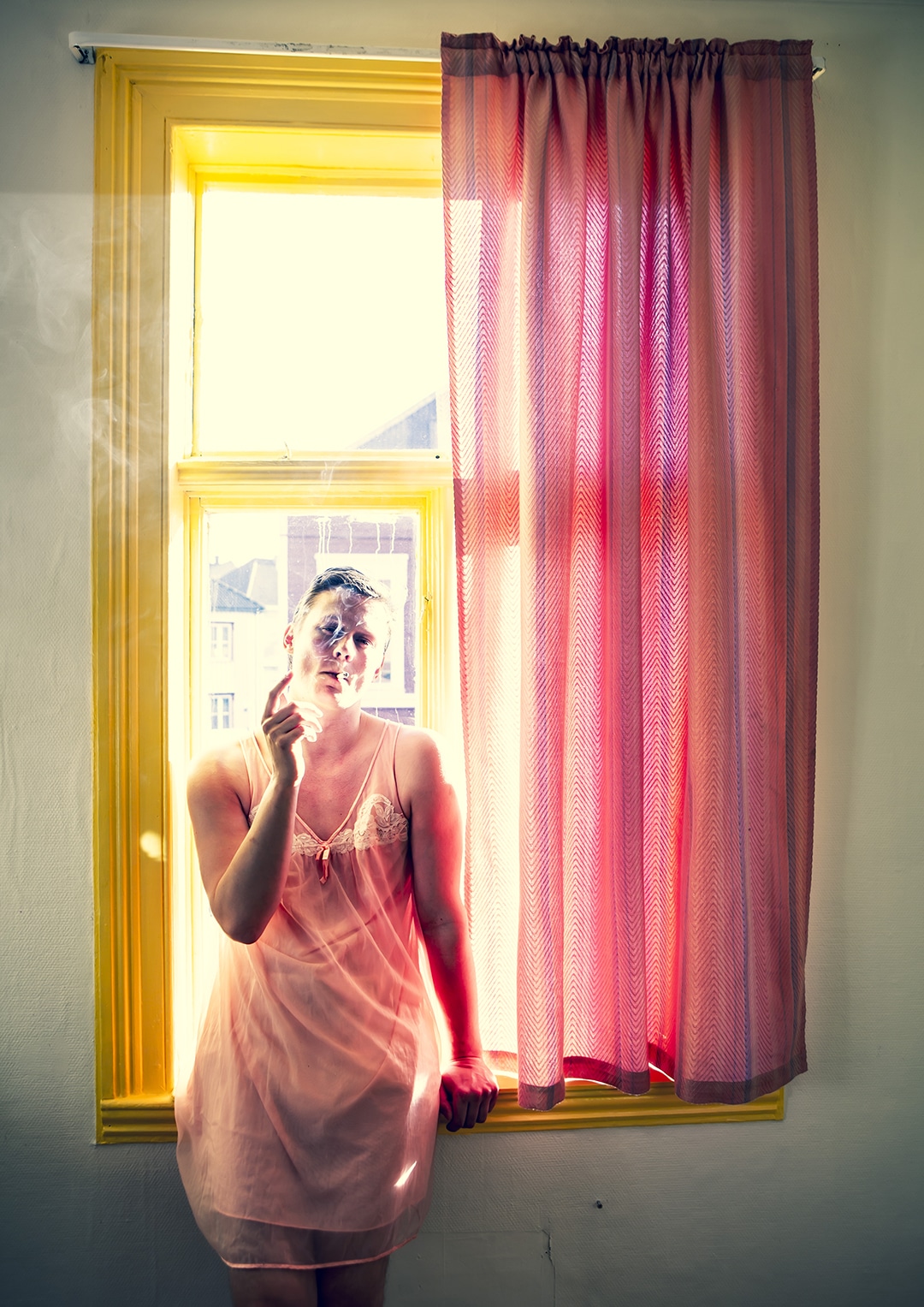 Fotografi: Bildet viser en ung man som røyker i rosa nattkjole foran et vindu.