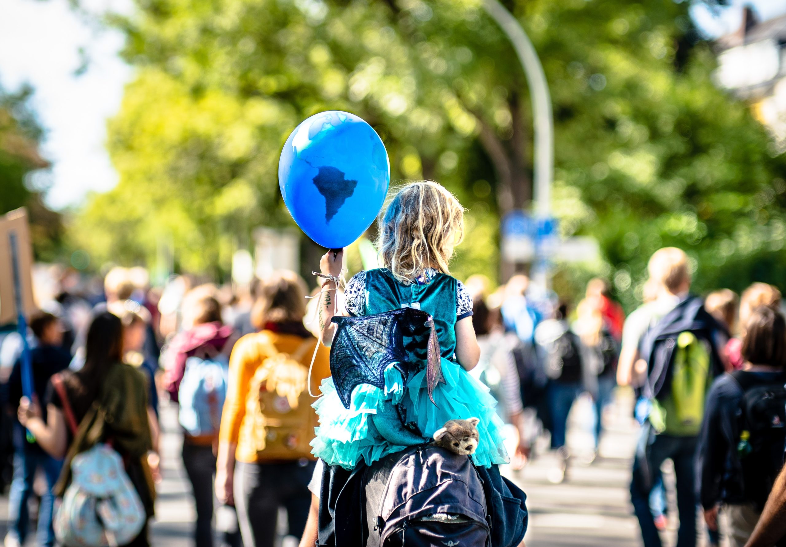 Fotografi: Bildet viser en ung jente som sitter på skuldrene til en voksen person, men en ballong i hånden. I bakgrunnen skimtes et demonstrasjonstog for klimaet.
